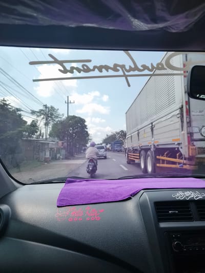 The image is taken from the interior of a car, looking forward through the windshield. Outside, the scene is a street with moderate traffic under a cloudy sky. On the dashboard, a purple microfiber cloth is placed near the windshield, and a pink "Hello Kitty" decal is visible on the textured dashboard surface. A calligraphic decal, possibly Arabic script, is present on the right side of the dashboard, above an air vent and a car stereo.

On the road, a motorcycle with a rider wearing a light-colored top and purple headscarf is moving away from the camera. To the right, a large, silver, box-shaped truck is driving in the same direction. Other vehicles, including cars and trucks, are visible further down the road, suggesting traffic flow. The street is lined with modest buildings and trees on the left. Power lines crisscross the sky, indicating an urban or semi-urban environment. The overall impression is a view of everyday traffic in a city, with personal decorations inside the car. The location is identified as Sidorejo, East Java.