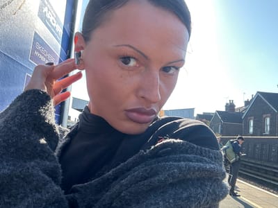 A bright outdoor selfie captures a woman from a slightly low angle, positioned in the foreground. She has light brown skin with visible freckles, dark hair, and is wearing eyeliner and reddish-pink lip color. Her eyes are light-colored, and she looks directly towards the camera with a neutral expression. She wears a textured dark grey cardigan or jacket over a black top. A metallic earring or earbud is visible in her right ear, and her fingers gently touch her earlobe.
The background reveals a sunny day with a clear blue sky. On the right, a large blue sign is partially visible, displaying white text that reads "SKECHERS," "HYPER B," "ARCH FIT," and "Product Certified By Experts." To the left, a train platform or paved area stretches into the distance, where a person, possibly a man or boy, is walking away from the camera, carrying a khaki or light green backpack. Further back, several buildings with dark roofs and windows are visible, suggesting an urban or suburban setting. The bright sunlight casts strong shadows and highlights across the scene, indicating a mid-morning or afternoon time of day.