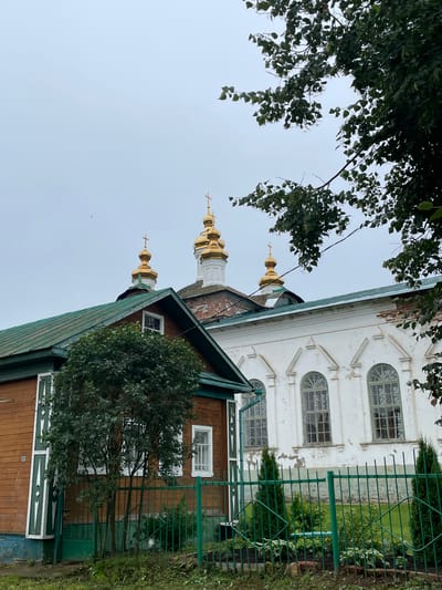 The image depicts a view of the Holy Trinity Church in the city of Kstinin, Russia, on a cloudy day. In the foreground, a low wooden building with a green roof is visible, featuring brown wooden siding and green decorative shutters. A green metal fence runs along the front of this building and extends towards a white building behind it. The white building has arched windows with green panes and shows signs of wear, with chipped paint and exposed brick in some areas. Behind the white building, the golden domes of the Holy Trinity Church rise, topped with crosses. Lush green trees and foliage frame the scene, with some branches extending into the upper portion of the image. The sky is overcast, contributing to a muted color palette. No people are present in the image.