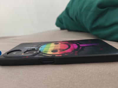 A smartphone with a colorful smiley face design is lying on a textured surface. The phone is black with a case that features a rainbow-colored smiley face with headphones. The camera lenses are visible on the back of the phone. The phone is angled, showing its side profile. In the background, a crumpled green fabric is visible, suggesting it might be a blanket or clothing. The overall scene is a close-up shot of a mobile device.