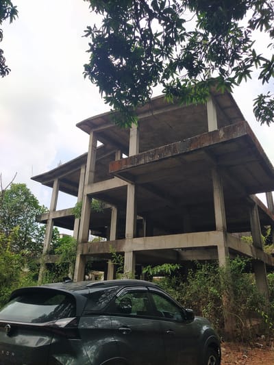 The image depicts the exterior of a multi-story, unfinished concrete building in Marna, India. The building's skeleton is exposed, revealing support beams and floor slabs. Vegetation, including vines and trees, grows around and through the structure. A dark-colored vehicle is parked adjacent to the building. The sky is visible through the incomplete structure and foliage.
