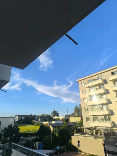 The image depicts a view from a balcony overlooking a suburban area in Katowice, Poland. In the foreground, a dark grey overhang from the balcony partially obscures the scene. The sky is a bright blue with wispy white clouds. To the right, a multi-story residential building with beige walls and numerous balconies is visible. Sunlight reflects brightly off the windows and balconies of this building. In the middle ground, there is a patch of bright green grass, surrounded by trees and other residential buildings. Further in the distance, power lines and a distant smokestack are visible. On the ground level, there are trash bins, a parked white van, and a parked black car.