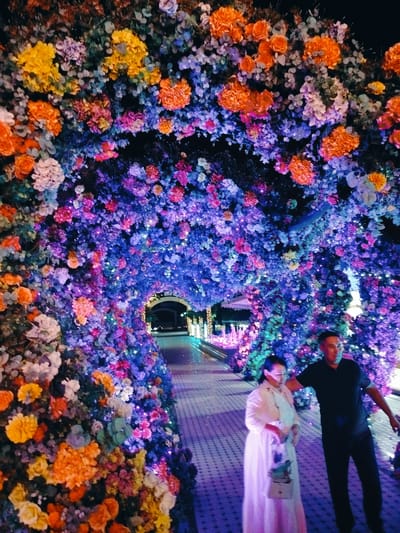 This media file captures a romantic scene within a vibrant floral archway illuminated by colorful lights, set in Aksuukent, Kazakhstan. The archway, densely packed with an array of artificial flowers in shades of orange, yellow, purple, and pink, creates a magical tunnel. Tiny fairy lights are interspersed throughout the foliage, casting a soft, enchanting glow.
The focus of the image is a couple walking through the archway. A woman in a flowing white dress and a light-colored shawl is on the left, carrying a stylish handbag. Her expression suggests a serene or perhaps contemplative mood. Beside her, a man in a black polo shirt and dark pants walks with his arm extended slightly behind her, indicating a sense of togetherness. His gaze is directed away from the camera.
The ground beneath them is paved with light-colored tiles arranged in a repeating pattern, reflecting the colorful lights from the floral arch. In the background, glimpses of a pathway and illuminated structures suggest an event or a decorated public space. The time of day appears to be evening or night, given the darkness of the sky and the prominence of the artificial lighting. The overall atmosphere is one of enchantment, romance, and celebration. There is no visible text in the image.