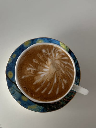 A top-down forensic image depicts a white ceramic coffee cup filled with a light brown beverage topped with elaborate white latte art, resembling a stylized explosion or starburst pattern, centered in the liquid. The cup's white handle is visible on the right. The cup rests on a ceramic saucer adorned with a reproduction of Vincent van Gogh's "The Starry Night," featuring swirling dark blue and yellow celestial patterns and a dark, tree-like element on the lower right, bordered by a thin gold rim. Both items are situated on a smooth, unadorned light grey surface. The scene appears to be an indoor setting, observed within the city of Kernen im Remstal, Germany. No individuals, overt actions, or interactions are present.