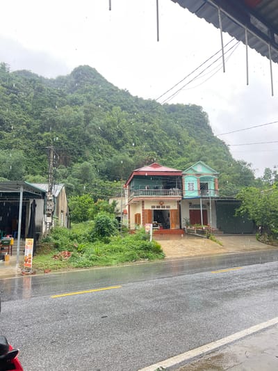 The image depicts a street scene in Sơn Dương Commune, Vietnam, during a rain shower. The foreground shows a wet asphalt road with a yellow line marking its center. In the middle ground, several buildings are visible. To the left, a covered structure with an open front and a sign is partially visible. Adjacent to it is a light-colored building with electrical conduits on its exterior. To the right, two multi-story houses stand side-by-side. The house on the left has a red roof and a balcony. The house on the right is painted in light blue and mint green colors, with a balcony featuring arched supports and a white window. Both houses have visible entrances and some potted plants. A lush, green hillside covered in dense vegetation rises behind the buildings, indicating a rural or semi-rural mountainous environment. The sky is overcast, and raindrops are visible in the air, suggesting active precipitation. The overall atmosphere is wet and somewhat subdued due to the weather.