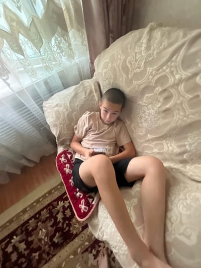 The media file shows a young boy lying on a bed. The boy is wearing a light-colored t-shirt and black shorts. He has a buzz cut and appears to be looking at the camera. His legs are bent, and one arm is resting on his stomach. The bed is covered with a textured, light-colored blanket. To the boy's right, there is a small, rectangular red cushion with white floral embroidery. Behind him, sheer white curtains with an embossed pattern are visible, suggesting a window. The overall lighting suggests it is daytime. The scene appears to be indoors, possibly a bedroom. The boy's expression is neutral. There is no visible text in the image. The location context provided is Leñgir, Kazakhstan.