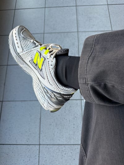 A person's left leg and foot, wearing a grey, washed denim pant and a New Balance 1906R sneaker, is captured in a medium close-up shot. The sneaker is predominantly white with silver accents and bright neon yellow "N" logos on the side. The foot is positioned at a slight upward angle, resting on a tiled floor that appears to be outdoors or in a well-lit indoor space with a blue-grey, speckled tile pattern. The background is blurred, but the distinct tiling suggests a public or semi-public area, potentially a plaza or a walkway in Badhoevedorp, Netherlands, given the context. The lighting is bright, casting subtle shadows and highlighting the textures of the denim and the sneaker's mesh and synthetic materials.