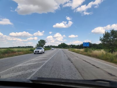 The media file shows a view from inside a vehicle driving on a paved road on a sunny day with scattered clouds. The road curves to the right and is bordered by green fields and trees. There are at least three cars visible on the road, including a white SUV in the foreground and two white cars further ahead. A blue directional signpost is visible on the right side of the road, indicating "Ruse" with an upward arrow and "Grivitsa" and "Dalen" with a rightward arrow. The scene suggests a journey through a rural area in Bulgaria. The time of day appears to be daytime, likely mid-morning or afternoon, given the brightness of the sky and the shadows. The weather is pleasant, with a clear blue sky and fluffy white clouds. The overall mood is calm and perhaps indicative of travel or transit.