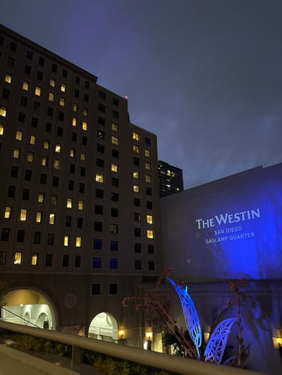 A low-angle shot reveals the Westin San Diego Gaslamp Quarter building at night, its name illuminated in blue. The building features many illuminated windows. To the right, decorative metal flora is subtly lit with blue, while arched openings show brightly lit interior spaces. A taller, modern building is visible behind the Westin. The evening sky is overcast.