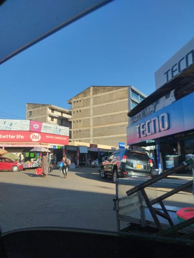 This street scene, captured during a bright, sunny day in Sheywe ward, Kenya, depicts a commercial area with buildings, shops, and vehicles under a clear blue sky.
In the midground, several people are present on the street. Two women walk towards the left, one carrying a red bag. A red car is parked on the left side of the road, while a dark SUV, appearing to be a Toyota Prado, is parked on the right. The image is taken from a slightly elevated perspective, possibly from inside a vehicle, with parts of the car's interior visible in the foreground.
Shopfronts line the street, featuring various mobile phone and electronics brands. Visible text includes a prominent red and white sign on the left for "better life" and "itel", alongside "MACOR COMMUNICATION". Further down, a "SAMSUNG" sign is noticeable. On the right, a large blue billboard for "TECNO" stands out, with "CITY WAY" and "Infinix" also legible. Other texts like "CITY 100" and "TELI" are also seen.
In the background, multi-story buildings with a uniform, unpainted concrete finish rise against the horizon. The strong daylight casts clear shadows, indicating either morning or late afternoon. The scene conveys the ordinary activity of a local commercial hub.