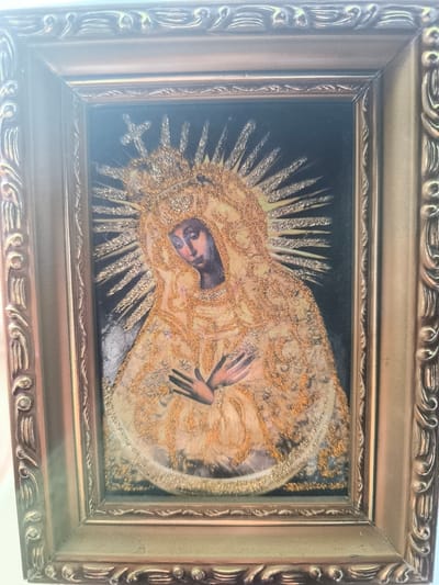 This is a framed religious painting of the Virgin Mary, likely depicting Our Lady of Guadalupe, given the iconography. She is shown with a golden crown and a halo of radiant rays. Her face is downcast, and her hands are crossed over her chest, conveying a sense of sorrow or solemnity. The artwork is highly ornate, with a rich golden embroidered robe featuring intricate patterns and shimmering embellishments. The background is dark, making the golden elements of the painting stand out. The frame itself is also elaborate, with a textured, antique gold finish and decorative carvings. The overall impression is one of reverence and artistic richness. There is no visible text in the image, nor are there any specific cues about the time of day, weather, or location beyond the general context of Almere, Netherlands, where the image was taken. The activity is static, being a painting rather than a depiction of an event.