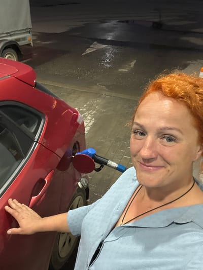 This media file captures a medium shot of a woman with vibrant red hair, leaning towards the camera and smiling. She is wearing a light blue, textured button-up shirt and a dark necklace. Her left hand is resting on the door handle of a red car, which is positioned in front of her.
The scene appears to be a car wash, indicated by the wet, reflective flooring and the presence of a gas nozzle inserted into the car's fuel tank. The red car has a dark interior visible through the window. The background is dark and somewhat indistinct, suggesting an indoor or covered area, possibly a bay at a car wash.
The lighting suggests it could be daytime, or a well-lit indoor environment. The woman's expression conveys a sense of lightheartedness and engagement with the camera. The context provided places the location in Можга, Russia. No visible text is present in the image.