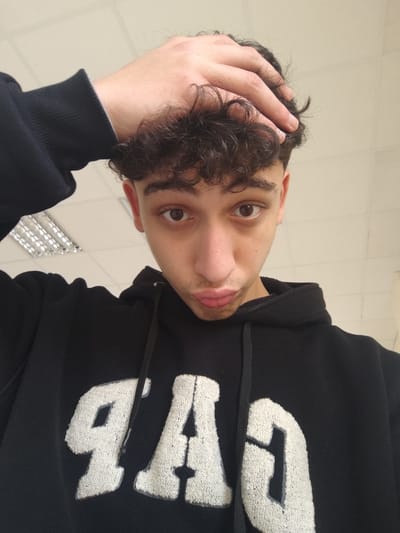 The image displays a young male subject in an indoor environment, located in Jibb as-Safa, Syria. The subject is oriented towards the camera, maintaining direct eye contact. He possesses dark, curly hair, which he holds with his right hand placed on the top of his head, fingers intertwined in the curls. His lips are noticeably pursed. The subject is wearing a black hooded sweatshirt, featuring the word "GAP" in large, white, textured applique letters across the chest. The background consists of a light-colored ceiling with visible grid lines and an overhead fluorescent light fixture containing multiple tubes, positioned in the upper left quadrant of the frame. The wall behind the subject is also light-colored.
