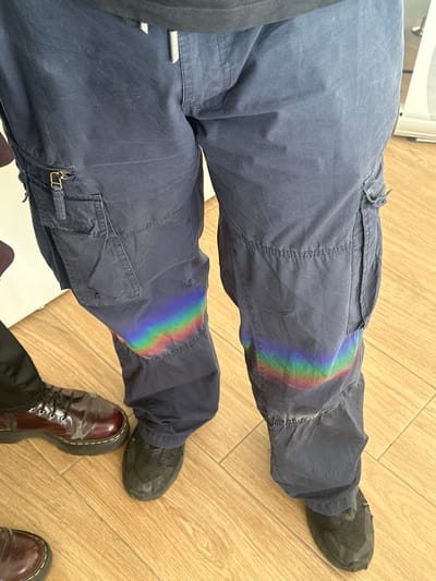 The photograph shows two pairs of legs clad in cargo pants and shoes, positioned on a light brown, wood-like floor. The top pair of legs wears navy blue cargo pants and dark gray shoes. A rainbow-like refraction of light is visible on the left leg of the top pair of pants. The bottom pair of legs wears navy blue cargo pants and burgundy, patent leather Doc Martens. The location appears to be indoors. There is no indication this image was taken in Quart de Poblet, Spain.