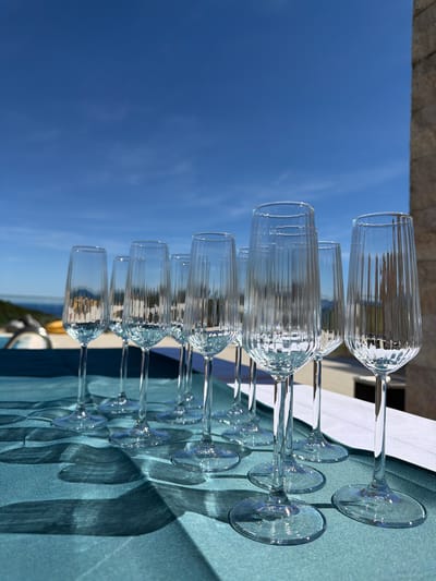 This image depicts a row of empty, clear fluted champagne glasses arranged on a teal-colored tablecloth outdoors. The glasses are tall and slender with ridged detailing on the bowls and clear, thin stems. Behind the glasses, a bright blue sky with a few wispy clouds is visible. To the right, a portion of a stone building or wall is present. The setting appears to be a terrace or balcony overlooking a body of water, likely the Adriatic Sea, given the context of Krimovice, Montenegro. The lighting suggests a sunny day, casting shadows of the glasses onto the tablecloth. There are no people or any liquid in the glasses. The environment is clean and tidy, suggesting a prepared setting for an event.