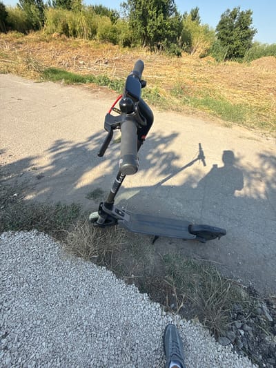 An electric scooter is parked on a paved path next to a dry, grassy area. The scooter is dark gray with a black deck and handlebars. A digital display is visible on the handlebars. The path is bordered by a gravelly area on the left and dry vegetation on the right. In the background, there are trees and a pile of dirt. The sun is casting long shadows from the scooter and a person, suggesting it is either early morning or late afternoon. The location is identified as the city of Pobeda, Bulgaria.