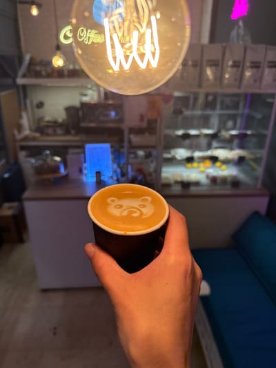 An overhead shot captures a person's hand holding a black paper cup of coffee with latte art of a bear face. The coffee cup is the closest object to the camera. Behind the cup, a blurred background depicts a cafe interior. A large, glowing filament light bulb hangs from the ceiling, dominating the upper portion of the image. Reflections of "Coffee" and a light bulb are visible on the bulb's surface. In the middle ground, a counter with a glass display case filled with baked goods is visible. To the right, a dark blue upholstered bench is present. The background also shows shelves stocked with coffee bags and a blue illuminated sign. The scene is set in a cafe in Veshki, Russia.