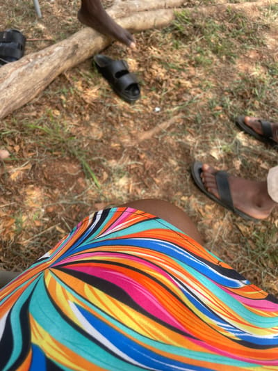 The image shows a ground-level view of a grassy area in Hayin Banki, Nigeria. Visible are several pairs of dark-colored sandals, some being worn and others lying on the ground next to a piece of wood. A person's leg and the edge of a garment with a bright, multicolored striped pattern are also visible. The ground consists of grass, dirt, and fallen leaves.