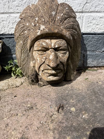 The image captures a weathered, carved stone ornament set outdoors, likely within a garden or patio in Longden, United Kingdom. The central subject is a stylized, abstract face or mask, possibly representing a grotesque or a Green Man motif, surrounded by intricate, radiating patterns that resemble deeply veined leaves or feathers. The stone carving itself is rough and textured, showing signs of age with discoloration and patches of white material, possibly lichen or weathered paint.
To the left and slightly behind the stone carving is a white, textured wall, which appears to be painted brick or stucco. Strong, directional sunlight illuminates the scene, casting sharp shadows and highlighting the textures of the stone, wall, and ground, indicating a bright daytime setting, likely morning or afternoon. In the foreground and to the right of the carving, there is a gray, rough concrete or paved surface, interspersed with small pebbles and bits of dirt. Small green plants are visible growing near the base of the stone carving and on the concrete, further emphasizing the outdoor environment. A tiny, dark insect can also be observed on the concrete surface. The overall impression is one of aged charm and natural integration into an outdoor space.