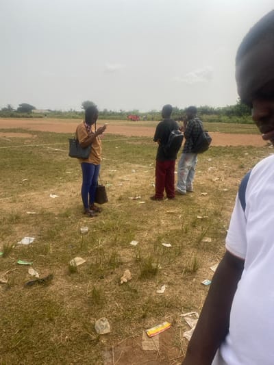 The image depicts an outdoor scene in Igbesa, Nigeria, under a bright, overcast sky. The ground is a mix of dry, sparse grass and reddish-brown dirt, heavily littered with various pieces of trash, including plastic wrappers and paper.
Four individuals are present in the foreground and midground. On the left, a woman with braided hair, wearing a tan long-sleeved top, blue trousers, and sandals, stands holding a white smartphone with both hands, looking down at it. She carries a dark patterned shoulder bag and a smaller dark patterned handbag. To her right, a man wearing a dark t-shirt, red trousers, and a black backpack stands with his back mostly to the camera, observing the distant background. Adjacent to him, another man wearing a dark plaid shirt, light-colored jeans with visible marks, and a black backpack also stands with his back partially to the camera, appearing to hold a phone to his right ear. In the extreme foreground on the right, the right shoulder and part of the head of a fourth individual, wearing a white t-shirt with a dark shoulder stripe, are visible, indicating their proximity to the camera.
In the far background, beyond a line of sparse vegetation, a dirt field is visible, where a group of individuals in red or orange clothing are gathered, suggesting an event or activity. A faint, light-colored line or rope extends horizontally across the midground on the dirt.
