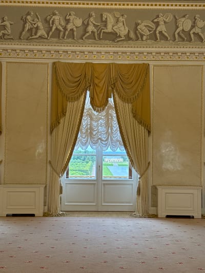 This image depicts a grand room, likely a ballroom or reception hall, within the Konstantinovsky Palace in Strelna, Russia. The scene is dominated by the luxurious interior, featuring a large window with elaborate gold and cream draperies, leading out to a view of a garden and a body of water with a distant building. The walls are adorned with classical relief sculptures above a decorative cornice. The floor is covered with a patterned carpet. On either side of the window, there are ornate white radiator covers. The overall impression is one of opulence and historical significance, consistent with a royal or governmental residence.