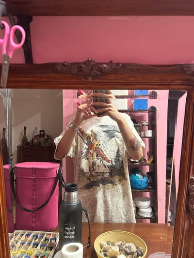A mirror reflects a person holding a phone, their face obscured. They wear a shirt with a wolf and eagle design. The mirror reflects a pink wall behind them. On the table in front of the mirror are a pink drum-shaped bag, a black thermos, a bowl of rocks, and a container of nail polish. Pink scissors sit at the mirror's edge. A wooden cabinet is visible in the mirror's reflection.