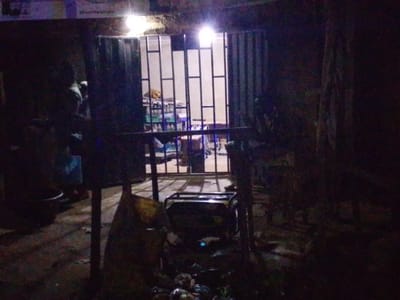 This image depicts the dimly lit exterior of a shop or business establishment in Kachia, Nigeria. The entrance is secured by a metal grate, allowing a partial view into the interior where shelves stocked with various items, possibly goods for sale, are visible. Overhead lighting illuminates the space. Outside the entrance, in the foreground, is a portable generator. The ground appears to be a dirt or unpaved surface, littered with debris and some discarded items, suggesting a somewhat unkempt outdoor area. The overall lighting suggests it is nighttime or a very dark, possibly overcast, day. The scene conveys a sense of basic commerce operating in a localized urban environment.