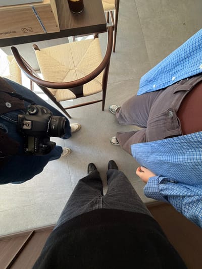 The image depicts an overhead view of the lower bodies and feet of four individuals gathered around a table in a café or interior setting within Marche-lez-Écaussinnes, Belgium. The floor is light grey tiled. To the immediate left, a person wears blue jeans and white sneakers, holding a black Canon DSLR camera on their lap. Directly opposite them, partially beneath a dark wooden table, is a vacant light-colored wooden chair with a woven seat. On the table, a dark glass containing a beverage (likely iced coffee) is visible, alongside a wooden box labeled "J.BOODLE LAB." To the top right, another person wears brown corduroy overalls over a dark top, a blue and white checkered shirt, and white sneakers. To the bottom right, the fourth person, presumably the photographer, wears black pants and black loafers. A third person's feet in white sneakers are visible at the far right. No direct interactions between the individuals are discernible.