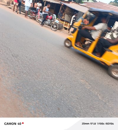 A blurred yellow auto rickshaw is captured in motion, moving from left to right. The driver, wearing a striped shirt, appears partially visible. In the background, people are standing by parked motorcycles near a building. The road occupies a significant portion of the foreground and extends towards the horizon. The photograph is taken in daylight with details of the street visible.