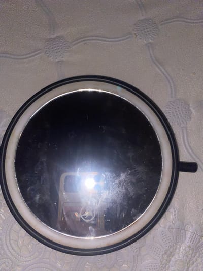 A close-up, overhead shot captures a round mirror with a black rim and a white inner ring, resting on a textured, light-colored surface that appears to be wallpaper or fabric. The mirror is slightly dusty, and a bright light source, likely a phone flash, is reflected in its center, obscuring much of the mirror's surface. The reflection also shows a portion of a mobile phone and a faint circular logo. The background surface exhibits a repeating floral or star-like embossed pattern. The overall scene is indoors, with no immediate indication of a specific city or location, but the prompt asks for a description in Афанасьево, Russia.