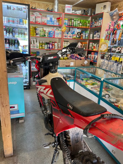 The image depicts an interior scene, possibly a small shop or convenience store, in Krasnyi Yar, Russia. In the foreground, a red and white dirt bike is parked, its rear section and seat clearly visible. The bike is covered in dirt, suggesting recent use. Behind the bike, shelves are stocked with various merchandise. To the left, a refrigerated display case, labeled "arnea" and "клап", contains bottled drinks. The shelves above and to the right display a mix of products, including cleaning supplies, toiletries, and potentially hardware or automotive parts. The overall environment appears to be functional and utilitarian, with wooden and plastic shelving units. No people are visible in the frame.
