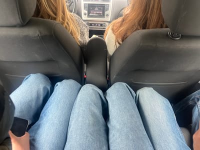 The image, taken from the back seat of a car, shows three people wearing blue jeans, with the two people in the front seats visible from the back of their heads and shoulders. A fourth arm, holding a cell phone, is seen on the left. The car's dashboard is visible in the background. It is not possible to ascertain the location of the image.