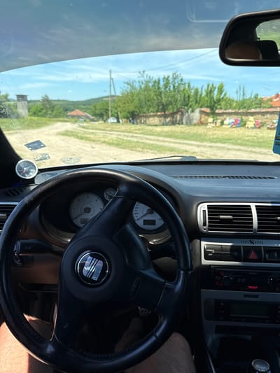 The image shows the interior of a car, specifically the dashboard and steering wheel. The steering wheel has a Seat logo. Through the windshield, a rural landscape is visible, potentially with a building and vegetation. The car appears to be in Goliam Dol, Bulgaria, based on visual context. A Sony car stereo is seen on the dashboard.