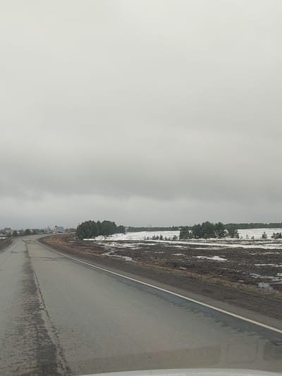 A photograph depicts a grey, overcast day in what appears to be a rural area. A paved road curves gently to the left, taking up the lower left of the frame. The road surface is wet and shows signs of wear, with visible cracks and patches. To the right of the road is a snow-dusted field, interspersed with patches of brown vegetation and scattered small trees. In the distance, a faint outline of structures can be observed near a treeline. The sky is uniformly grey and occupies the majority of the upper portion of the photograph, with no visible sun or breaks in the cloud cover. The setting is possibly near Povarenki, Russia, based on context.