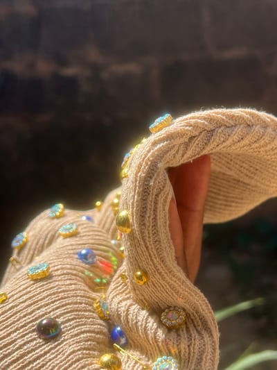 The image presents a close-up, high-detail view of a light-beige, ribbed knit garment, possibly a sleeve or a portion of a larger textile item. The fabric is heavily embellished with an assortment of decorative elements, including numerous gold-toned dome-shaped studs, various iridescent and faceted gemstones in gold settings (some displaying an aqua hue, others a rainbow spectrum), dark blue teardrop-shaped beads, and several gold-toned safety pins. A person's hand or wrist, characterized by dark-toned skin, is visible within or adjacent to the garment, suggesting the item is being worn or held. The garment is slightly creased and folded, indicating movement or a natural drape. The scene is illuminated by strong, direct sunlight, creating bright reflections on the metallic studs and iridescent gemstones. The background is out of focus and appears dark brown to black, with a blurred green element discernible in the lower right quadrant, suggesting an outdoor environment. This scene is located in Shen, Nigeria.