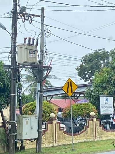 The image shows utility poles covered in wires, with electrical equipment attached. A yellow road sign with two horizontal black bars is visible. Beyond the poles are trees and a building. The building has a sign that reads "Bhayangkari." Green bushes and a red roof are visible, along with a partially obscured car. The sky is overcast.
