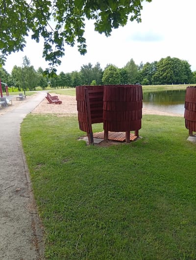 The image depicts an outdoor scene in Šaukėnai, Lithuania, near a body of water. In the foreground, a grassy area is visible, with a path to the left. Two cylindrical wooden structures, resembling changing stalls or outdoor showers, stand on the grass. These structures are made of vertical wooden slats and have wooden slatted floors. One structure is more fully visible than the other, which is partially cropped on the right side of the image. In the middle ground, a sandy area contains several red lounge chairs. Beyond the sandy area, a calm body of water reflects the sky and surrounding greenery. A line of trees borders the water and extends across the background, with a slightly overcast sky above. The overall environment suggests a recreational area, possibly a park or beach.