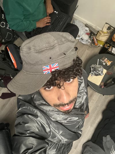 The image depicts an indoor scene in Marcoussis, France. In the foreground, a male individual with brown skin, dark curly hair, a mustache, and a short beard looks directly upward at the camera. He wears a grey/silver reflective puffer jacket and a grey cadet-style cap featuring a British Union Jack flag patch on the front brim, positioned over his forehead.

In the upper left portion of the frame, a second individual is partially visible from the chest down, seated on the floor. This person wears a green long-sleeved top and black trousers, and their hands are positioned on the keyboard of a black laptop. Adjacent to them is a black backpack with partial white lettering.

To the right, a round, dark-colored table holds various items: a pack of cigarettes with an orange elastic band, a crumpled silver foil packet, a light-colored rectangular object, scattered reddish-brown pellets, and a dark bottle. The floor is covered with light-toned laminate or wood and appears cluttered, particularly on the right side, with a white plastic bag, a silver insulated water bottle, a white mug, an orange and black box, and a patterned fabric container. A plain white wall forms the background.