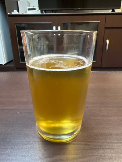 A clear glass filled with a golden-yellow beverage, likely beer, sits on a dark brown wooden surface. The beverage has a frothy head of white foam on top. The background is blurred, but appears to be an indoor setting with dark wooden cabinets and some electronics on a shelf. There is no indication of the specific city or country.