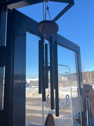 A dark, multi-tubular wind chime, suspended by natural fiber rope from an overhead fixture, hangs centered in the foreground. It consists of dark gray cylindrical chimes attached to a reddish-brown wooden disk, with a matching wooden clapper at the bottom. The wind chime is positioned within a dark-framed glass doorway or large window, looking out onto a bright, clear blue sky.
Through the glass, a winter or early spring landscape is visible in Сундур, Russia, featuring snow-covered ground with patches of brown earth, scattered bare trees, and distant low-rise buildings. Reflections on the glass overlay the exterior view, displaying Cyrillic text such as "БЕЗ ОБЕД" (without lunch break) and "ВХОД" (entrance), along with the numeral "8". A small white note, possibly a printed sign with Cyrillic text including "рассылка" and "до 12:00", is taped to the interior of the glass or a door handle, partially obscured by the wind chime. The partial reflection of an individual, primarily the right side of their head with light-colored hair and a portion of their upper body clad in a light blue garment, is visible on the right side of the glass, with their right hand positioned near their mouth.