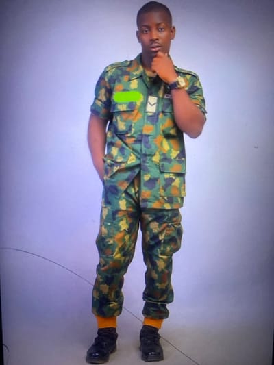 A young man in camouflage military fatigues stands with one hand near his mouth and the other resting on his side. He wears a wristwatch and a rank insignia on his upper left chest. His pants are also camouflage and are tucked into black boots over orange socks. The background is a plain grey. The image is rotated ninety degrees counterclockwise.