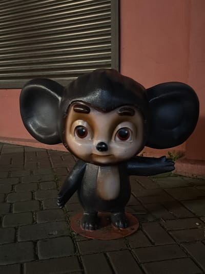 A statue of a cartoon character, Cheburashka, stands on a cobblestone pathway in front of a pink building. The statue is dark brown with large ears, a tan face, and black eyes and nose. It is wearing a light tan chest piece. The character's right arm is extended to the right. The building behind the statue has a dark, corrugated metal door or shutter. The scene appears to be outdoors, possibly in a public space. The location is identified as Kislovodsk, Russia.