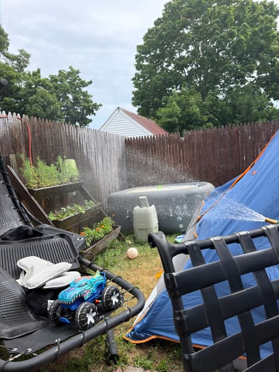 The image depicts an outdoor scene with a blue tent and an inflatable hot tub. A hose is spraying water onto the tent, creating a visible mist. In the foreground, a black mesh chair with a black woven seat is partially visible. Other objects in the scene include a blue toy, two white mesh insoles, and two small black and white wheels attached to a metal frame. A wooden fence and lush green trees form the backdrop, suggesting a backyard setting. A small portion of a white building with a triangular roof is visible behind the fence. The sky is overcast, indicating cloudy weather. The overall impression is that of an outdoor area being cleaned or prepared, possibly for leisure activities. The time of day appears to be daytime, given the natural light. The location is identified as Riverhead, United States of America. There is no visible text in the image.