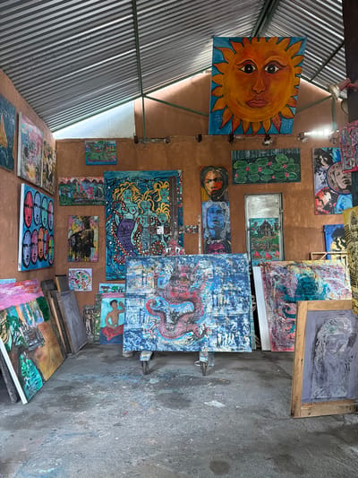 This image depicts an indoor art studio in Tejakula, Indonesia, filled with numerous paintings. The studio has a corrugated metal roof and earthen-colored walls. A large, vibrant painting of a sun with a human face is prominently displayed on the back wall. Other paintings on display include a large blue canvas featuring mythological figures, a detailed depiction of a red dragon, portraits, landscapes, and abstract pieces. Several canvases are propped up against walls or displayed on easels. The floor appears to be concrete, splattered with paint. There are no people visible in the image.