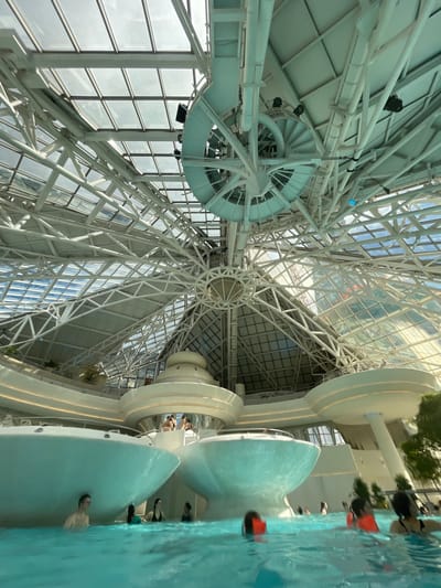 The image captures the interior of Caldea, a thermal spa located in Les Escaldes, Andorra. The focus is on the building's architecture, with a view looking upward towards the structural framework of the roof. The roof is a network of white steel beams and glass panels, creating a complex geometric pattern against the sky. A circular architectural element is seen on the left side, near the beams. On the right side of the image, parts of a swimming pool area are visible, with people swimming. The image is rotated, giving a tilted perspective of the scene.
