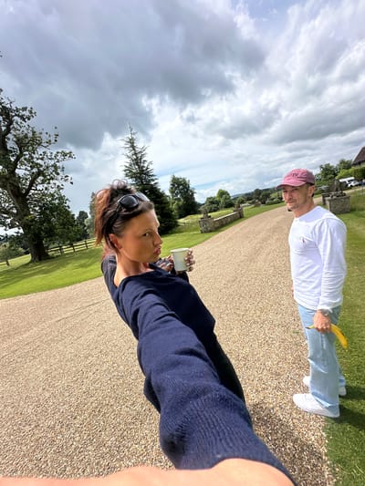 A selfie taken outdoors in Chichester, United Kingdom, shows two people on a gravel driveway. In the foreground, a woman is turned towards the camera, holding a white cup and making a pouting face. She is wearing a dark blue long-sleeved top and her hair is pulled up in a messy bun with sunglasses on top. Behind her and to the right, a man is looking at the camera. He is wearing a white long-sleeved t-shirt, light blue jeans, white sneakers, and a pink baseball cap. He is holding a yellow banana in his right hand. The background features a manicured lawn, trees, a wooden fence, and some stone structures. The sky is overcast with dramatic clouds.