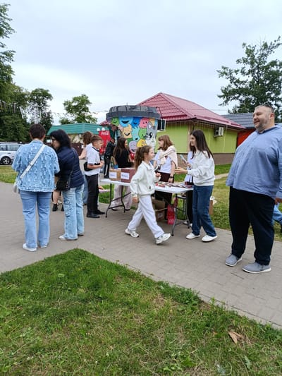 An outdoor scene in Smilyavitski Sel'ski Sovet, Belarus, features a gathering of people around a table laden with goods. A diverse group, including adults and children, are engaged in various activities. Some individuals are standing, observing, or interacting with items on display. A young girl in a white tracksuit is in motion, walking past the table. Another woman, dressed in a white hoodie and jeans, is holding a phone, possibly in the process of making a purchase or sharing information. A large man in a blue shirt and dark pants stands prominently in the foreground, looking to his right and smiling. Behind the table, several other individuals, including a young boy in a white shirt and dark pants, are present. The background reveals a building with a red roof, a vibrant, cartoon-decorated structure resembling a large trash can, and trees. The ground is a paved walkway bordered by grass. The overall atmosphere suggests a casual outdoor event or market.