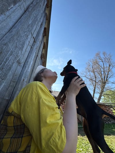 A person wearing a yellow shirt is holding a black dog up in the air. The dog has a red collar. The sky is blue and there is a bare tree in the background. A weathered wooden structure is partially visible. The location is Усть-Тунтор, Russia.
