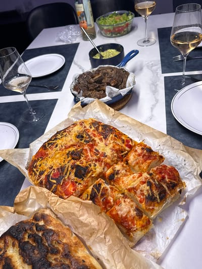 The image shows a dining table set for a meal. On the table, there are plates, wine glasses filled with white wine, a salad in a clear bowl, a blue bowl containing a green dip, a blue pot holding cooked meat, pizza on parchment paper, and flatbread on parchment paper. A bottle of wine is also visible. The table has a patterned black and white surface. The setting suggests an indoor dining environment. The location in the city of Ariel, Palestinian Territories cannot be determined from the provided image.