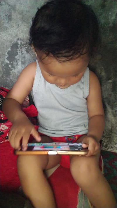 The image shows a young child sitting on a red and green patterned surface. The child is wearing a light grey tank top and red underwear with a watermelon pattern. They are looking down at a smartphone held horizontally in their hands, appearing to be interacting with it. The child's skin tone is dark, and they have dark, curly hair.
The background appears to be a rough, grey concrete wall. The lighting suggests it is daytime, possibly indoors or in a shaded outdoor area. The overall scene conveys a sense of quiet absorption as the child focuses on the screen. No text is visible in the image. The location is Reudeuep, Indonesia.